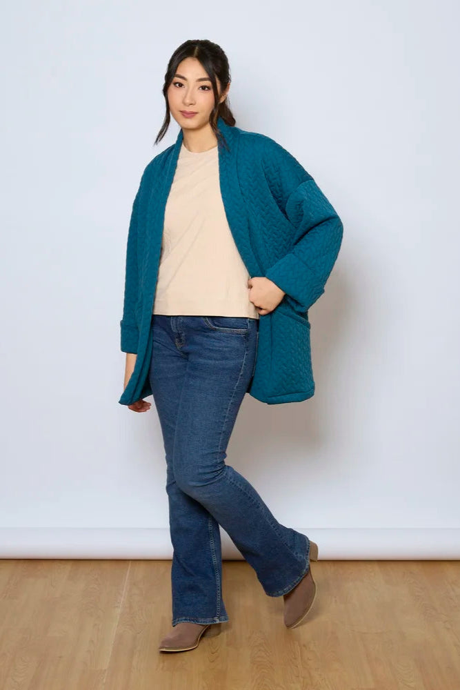A woman wearing the Ina Coatigan by Tangente in Peacock, an open front cardigan in a cable-knit textured fabric, with dropped sleeves and patch pockets. She is wearing it with a white shirt and jeans and standing in front of a white background. 