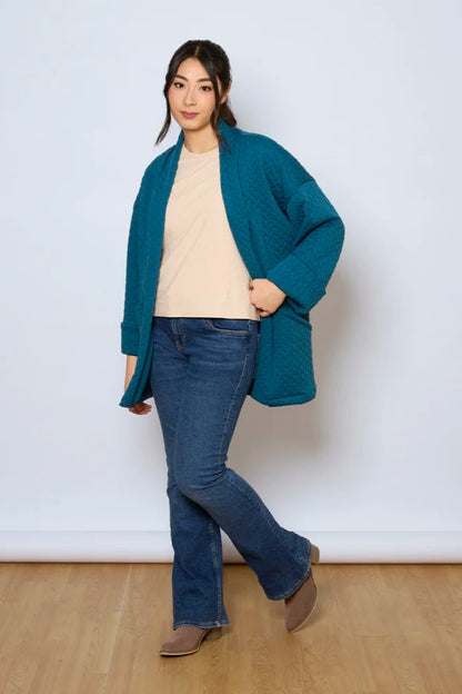 A woman wearing the Ina Coatigan by Tangente in Peacock, an open front cardigan in a cable-knit textured fabric, with dropped sleeves and patch pockets. She is wearing it with a white shirt and jeans and standing in front of a white background. 