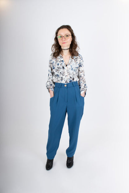 A woman wearing the Laura Pants by Infime in Blue, with a floral blouse, standing in front of a white background 