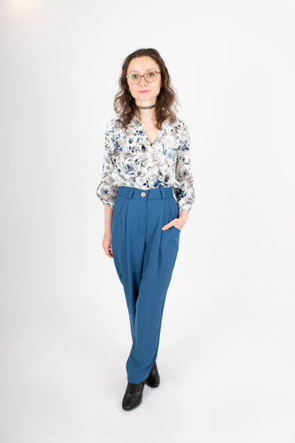 A woman wearing the Laura Pants by Infime in Blue, with a floral blouse, standing in front of a white background 