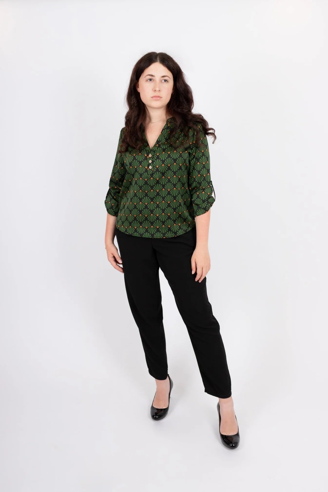 A women wearing the Leonie Blouse by Infime in Black with Green print, featuring a notched V-neckline, three button placket, and 3/4 sleeves. She is wearing it with black pants and standing in front of a white background. 