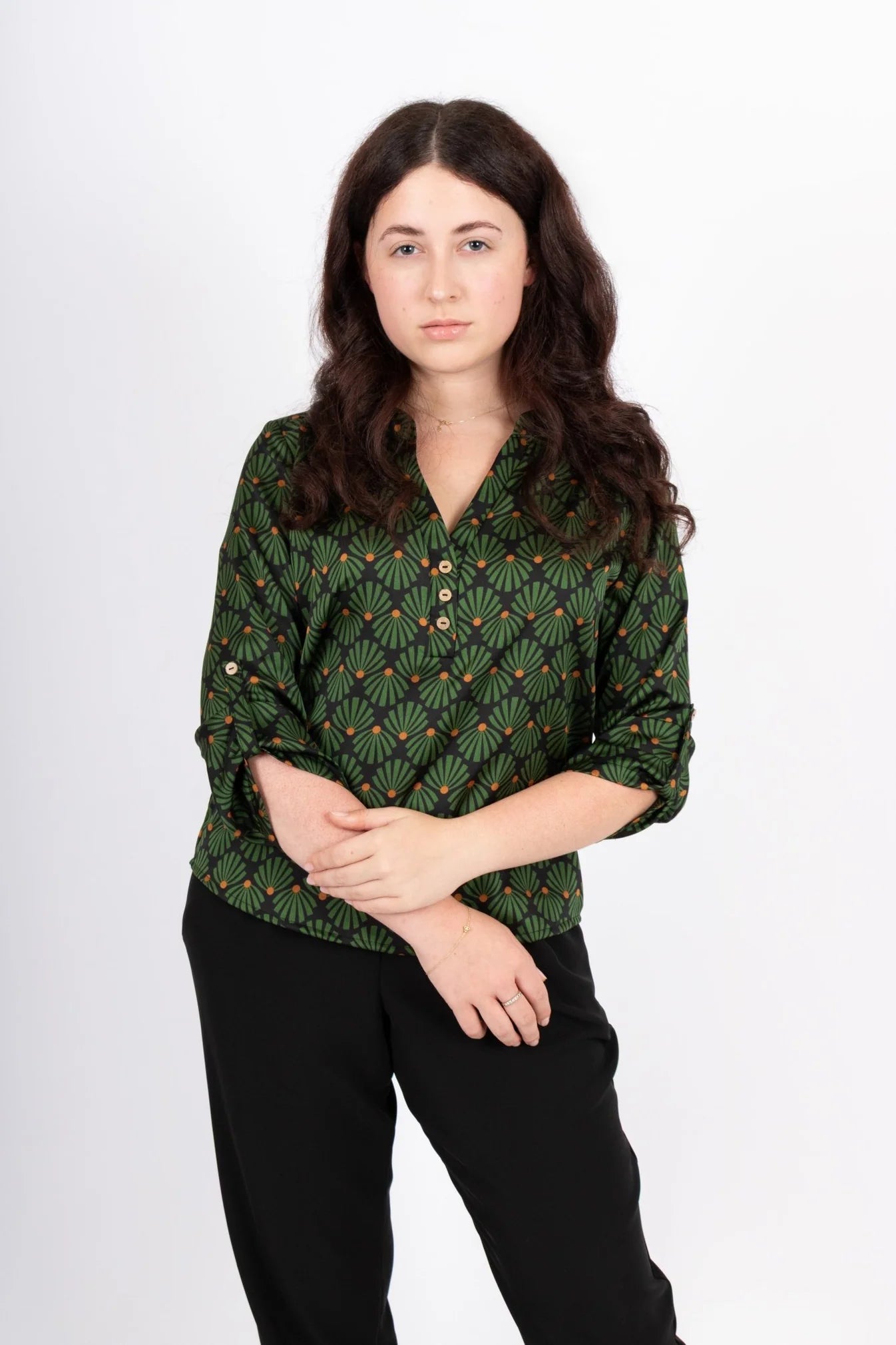 A woman wearing the Leonie Blouse by Infime in Black with Green print, featuring a notched V-neckline, three button placket, and 3/4 sleeves. She is wearing it with black pants and standing in front of a white background. 