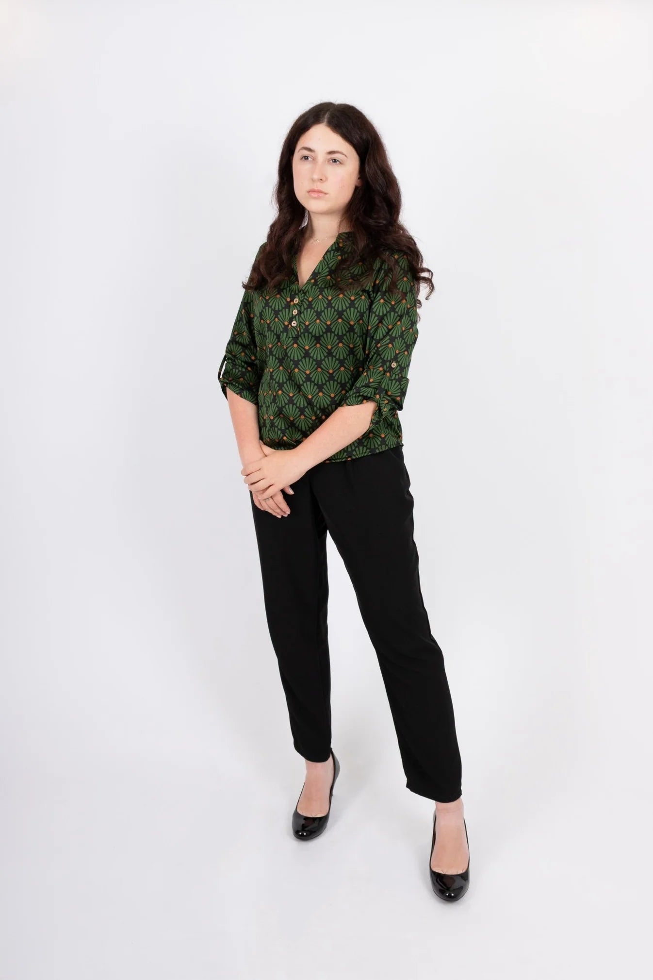 A woman wearing the Leonie Blouse by Infime in Black with Green print, featuring a notched V-neckline, three button placket, and 3/4 sleeves. She is wearing it with black pants and standing in front of a white background. 