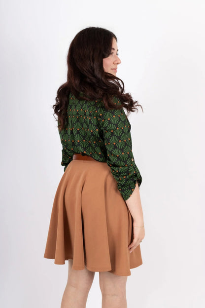 Back view of a woman wearing the Leonie Blouse by Infime in Black with Green print, featuring a notched V-neckline, three button placket, and 3/4 sleeves. She is wearing it with a tan skirt and standing in front of a white background. 