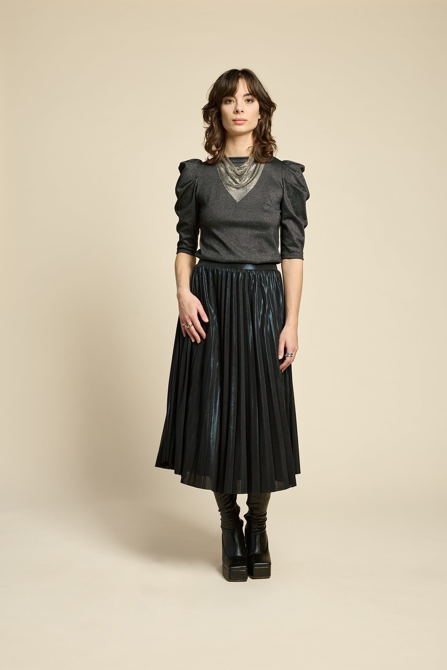 A woman wearing the Insomnie Top by Cokluch in Facet Black, a metallic piece with 3/4 puffed sleeves, darts at the bust, and a slim fit. She is wearing it with the Feyer Skirt in Glow Black Knit and standing in front of a beige background. 