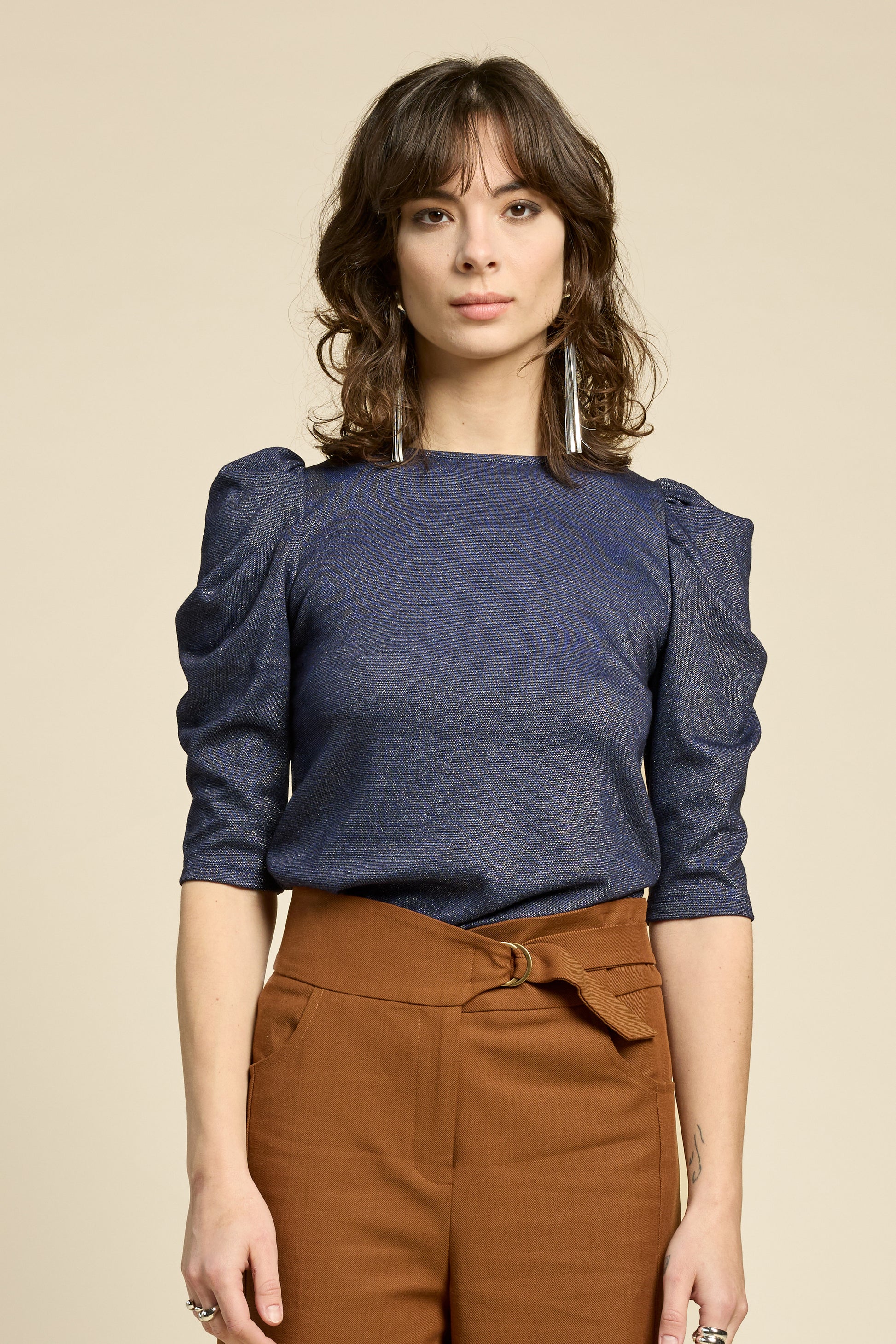 A woman wearing the Insomnie Top by Cokluch in Facet Twilight, a metallic piece with 3/4 puffed sleeves, darts at the bust, and a slim fit. She is wearing it with brown pants and standing in front of a beige background. 