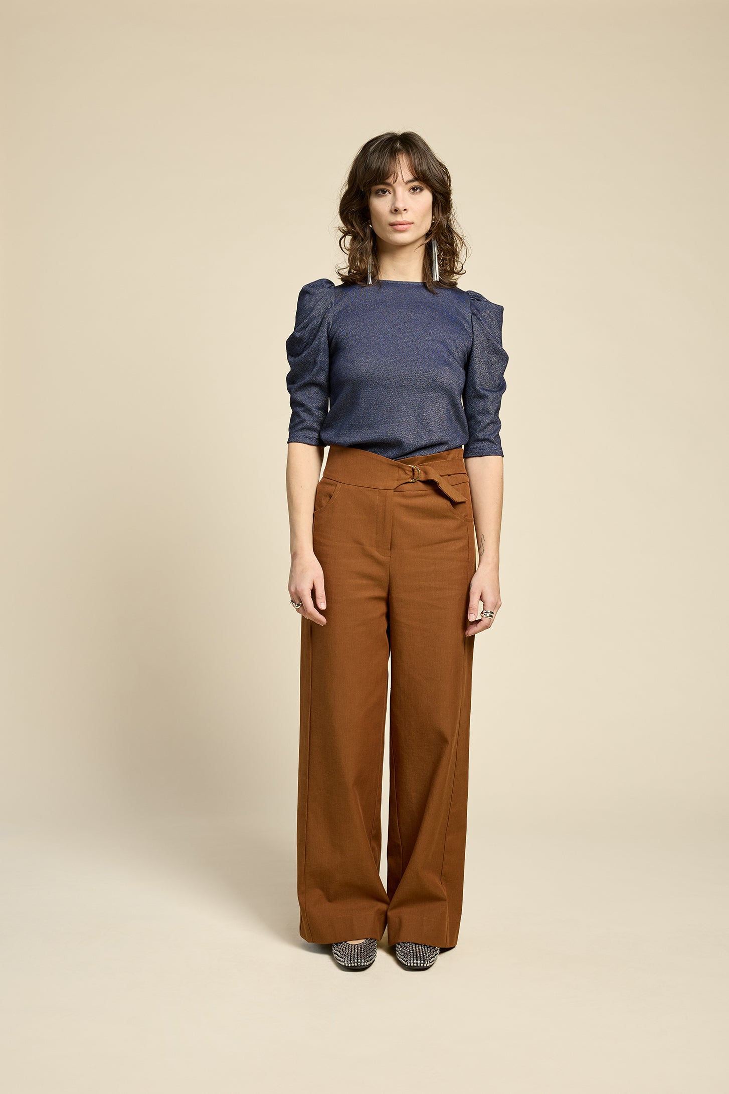 A woman wearing the Insomnie Top by Cokluch in Facet Twilight, a metallic piece with 3/4 puffed sleeves, darts at the bust, and a slim fit. She is wearing it with brown pants and standing in front of a beige background. 