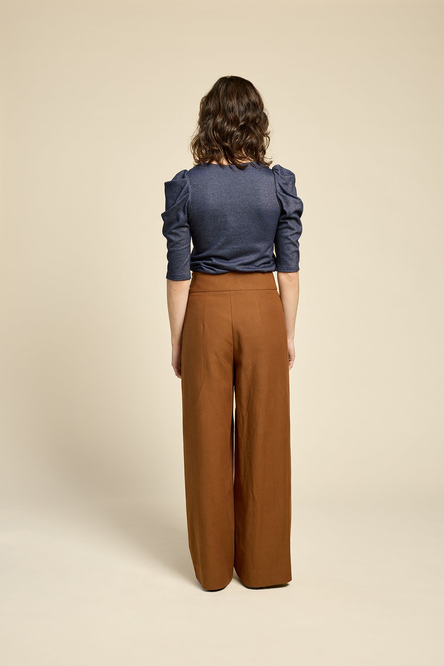 Back view of a woman wearing the Insomnie Top by Cokluch in Facet Twilight, a metallic piece with 3/4 puffed sleeves, darts at the bust, and a slim fit. She is wearing it with brown pants and standing in front of a beige background. 