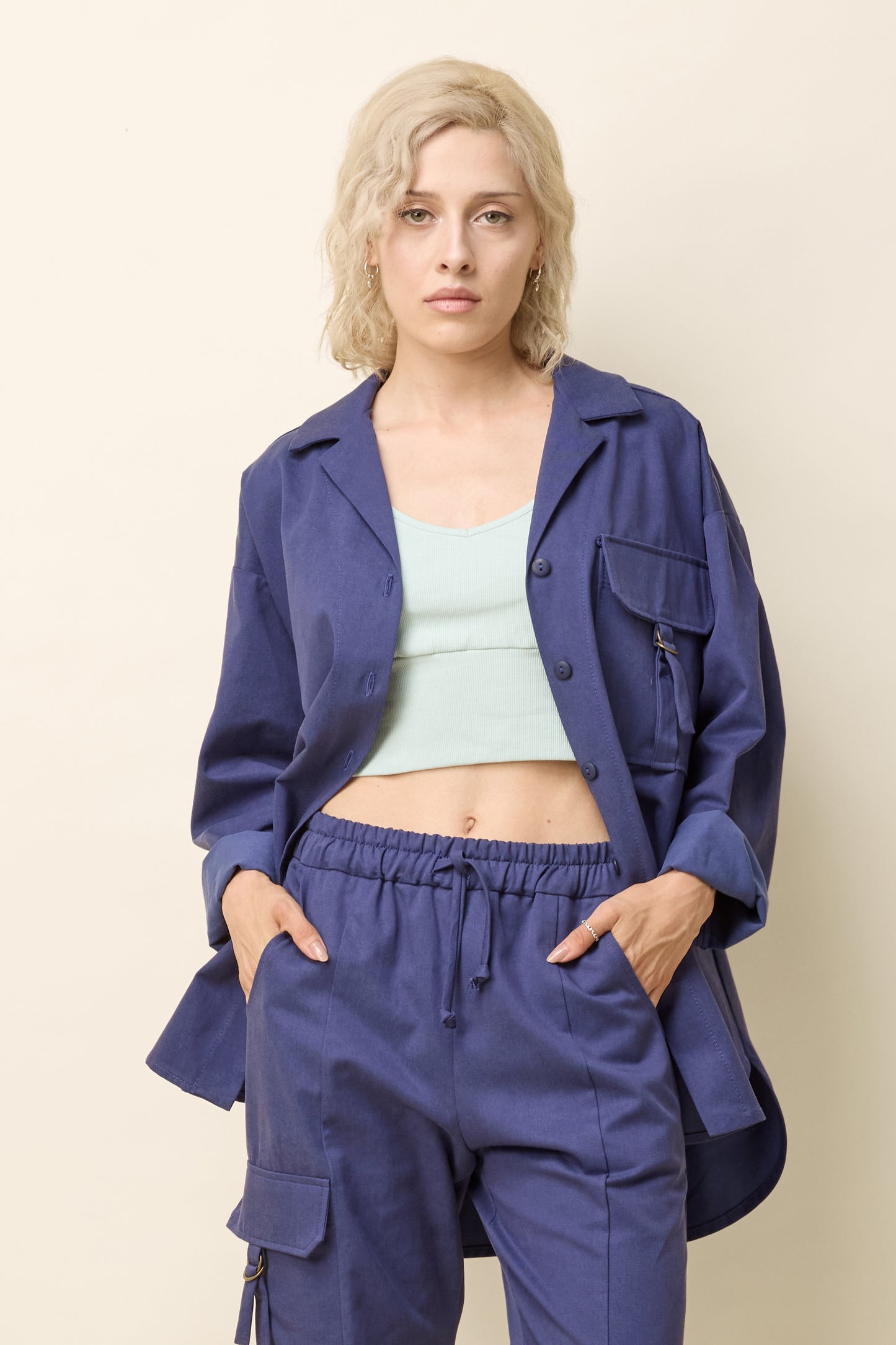 A woman wearing the Iyrna Shirt by Cokluch in Twiight blue, an oversized long sleeved button front shirt with chest flap pocket with a ring closure detail. She is wearing it with matching pants and standing in front of a beige background.  