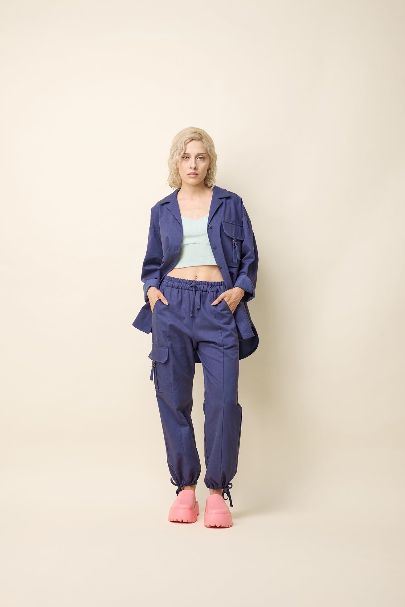 A woman wearing the Iyrna Shirt by Cokluch in Twiight blue, an oversized long sleeved button front shirt with chest flap pocket with a ring closure detail. She is wearing it with matching pants and standing in front of a beige background.  