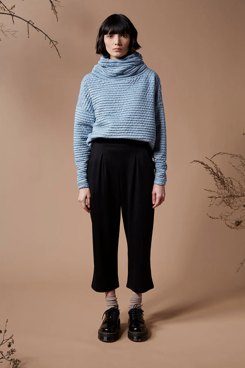 A woman wearing the Wilfred Pants by Melow in Black, with cropped legs, a pull on waist band and front pleats, with a blue sweater, standing in front of a beige background. 