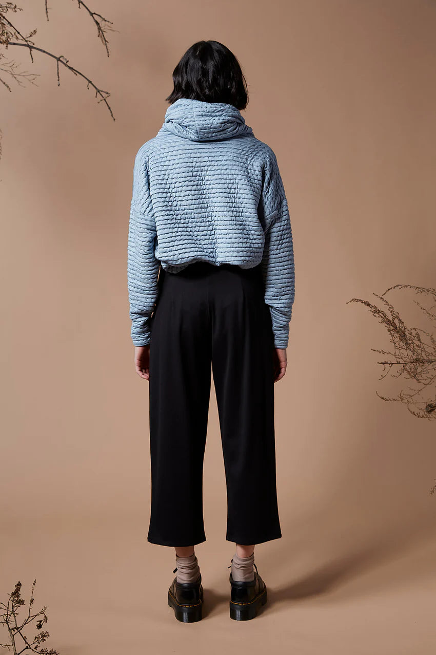 Back view of a woman wearing the Wilfred Pants by Melow in Black, with cropped legs, a pull on waist band and front pleats, with a blue sweater, standing in front of a beige background. 