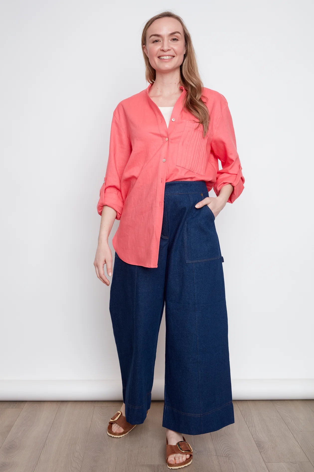 A woman wearing the jasmine Long Sleeve Shirt by Julei in Coral, a classic dress shirt style with a mandarin collar, pleated pocket, and cuffed sleeves. She is wearing it with jeans and standing in front of white background. 