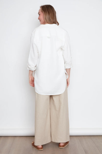 Back view of a woman wearing the jasmine Long Sleeve Shirt by Julei in Natural White, a classic dress shirt style with a mandarin collar, pleated pocket, and cuffed sleeves. She is wearing it with jeans and standing in front of white background. 