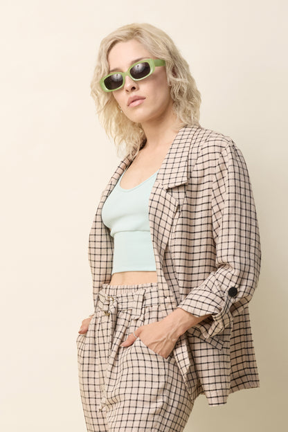 A woman wearing the Jennifer Jacket by Cokluch in Natural Utopia - a beige checkered pattern - with a wide lapel collar, two front buttons, angled front helm, and adjustable sleeves. She is wearing it with matching pants and standing in front of a beige background. 