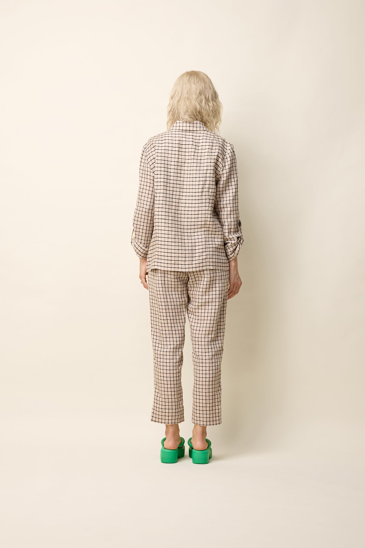 A back view of a woman wearing the Jennifer Jacket by Cokluch in Natural Utopia - a beige checkered pattern -  with a wide lapel collar, two front buttons, angled front helm, and adjustable sleeves. She is wearing it with matching pants and standing in front of a beige background. 