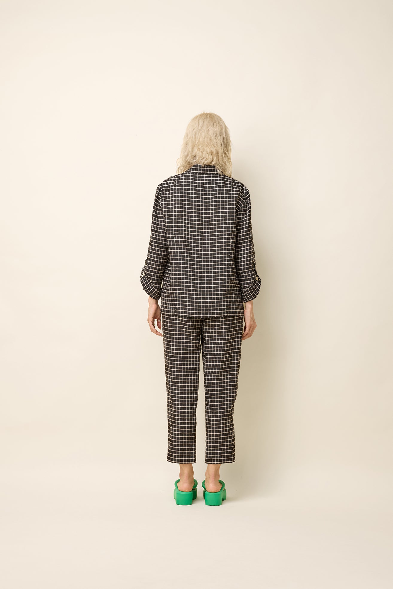 Back view of a woman wearing the Jennifer Jacket by Cokluch in Black Utopia - black checkered pattern - with a wide lapel collar, two front buttons, angled front helm, and adjustable sleeves. She is wearing it with the matching Yasmina Pants and standing in front of a beige background. 