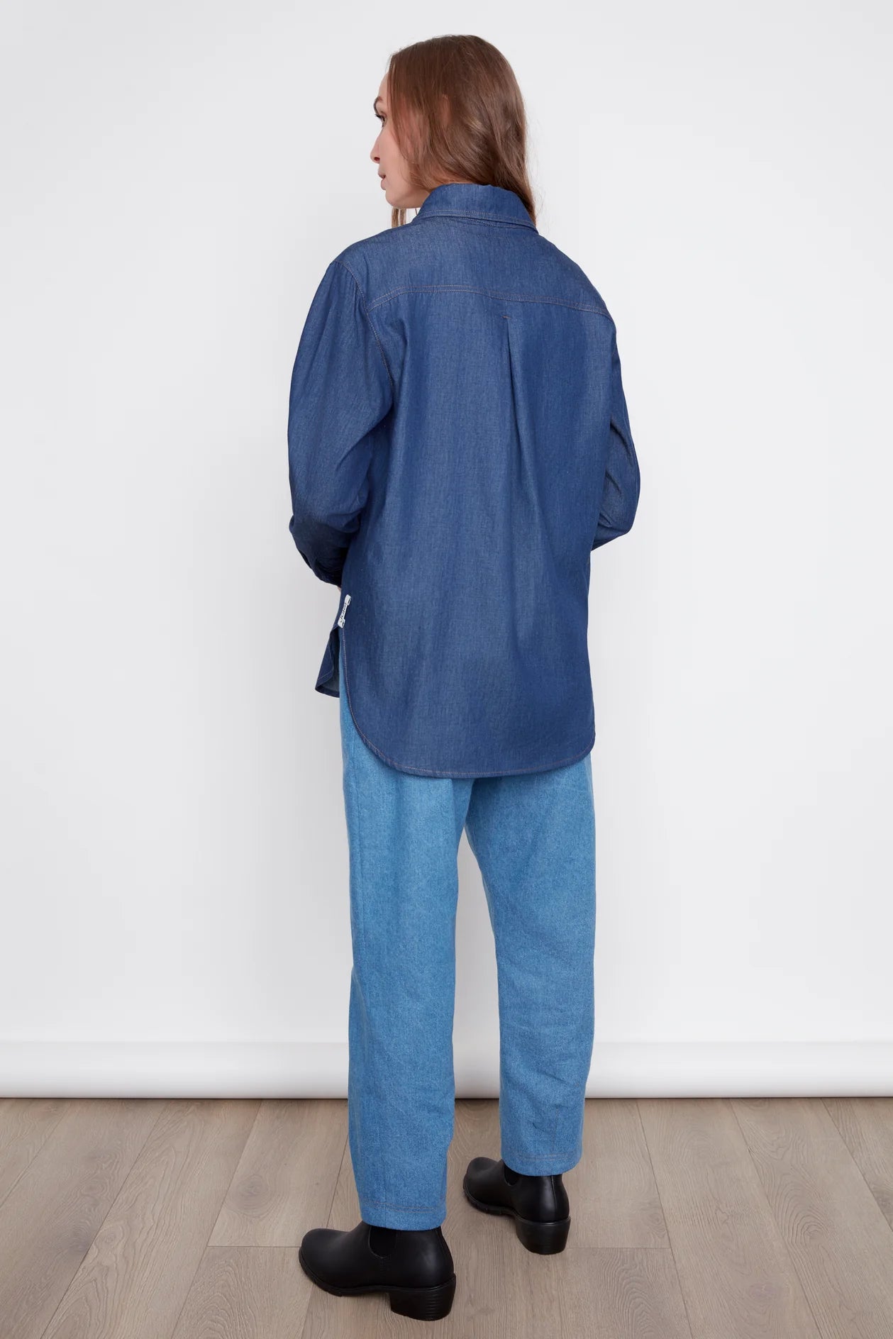 Back view of a woman wearing th eJasmine Denim Shirt by Julei, a boyfriend style button front shirt with a chest pocket and a large front patch pocket. She is wearing it with jeans and standing in front of a white background. 