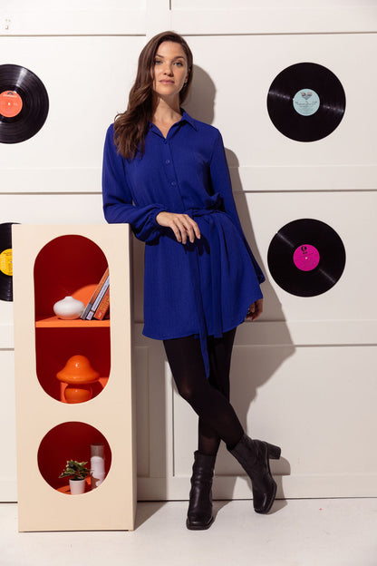 A woman wearing the Alanis Dress by Cherry Bobin in Blue, a classic shirt dress with a collar, button front, and tie belt and a rounded above the knee hemline. She is wearing it with black tights and is standing in front of a white wall with black vinyl records hanging on it. 