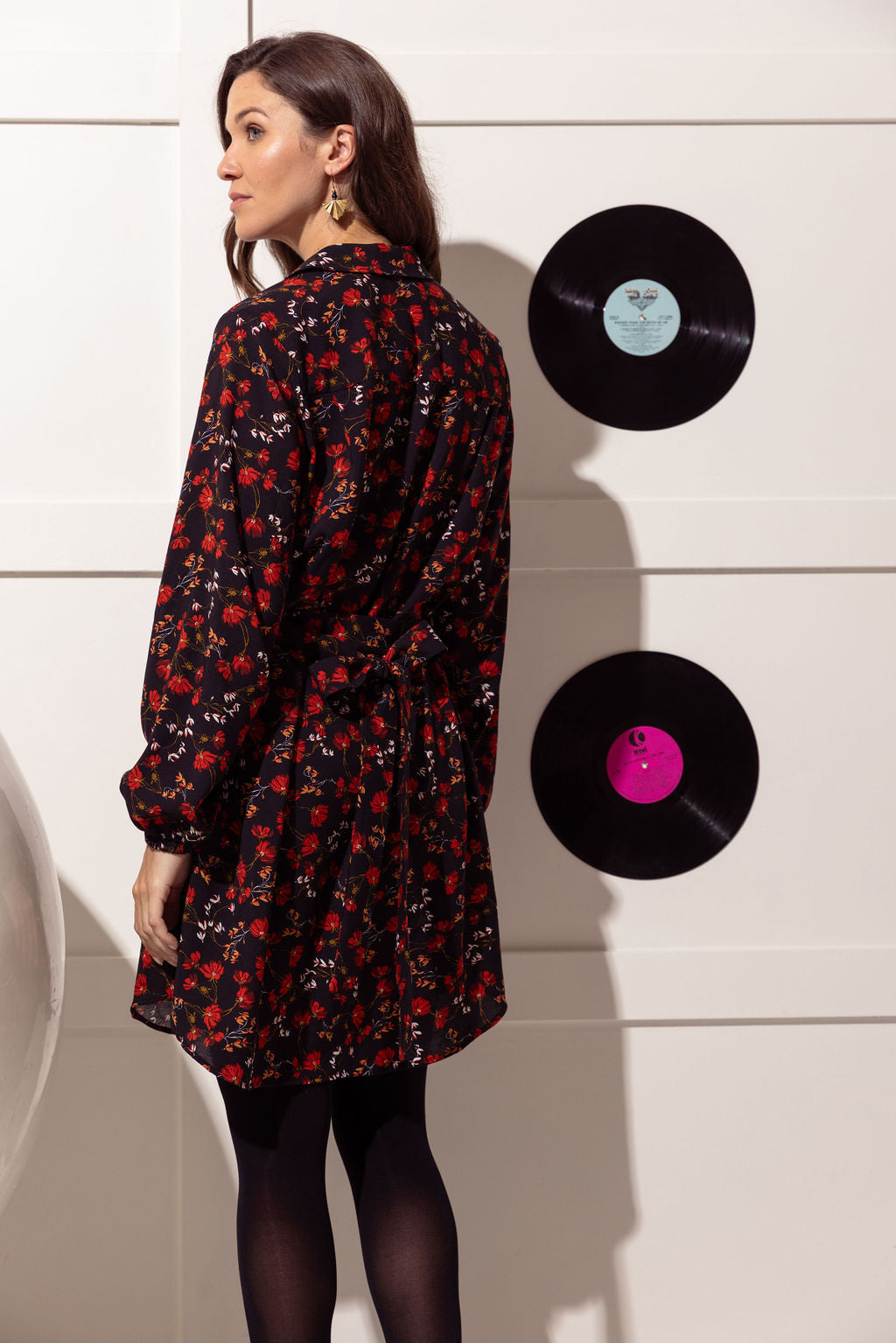 Back view of a woman wearing the Alanis Dress by Cherry Bobin in Black with Red Flowers, a classic shirt dress with a collar, button front, and tie belt and a rounded above the knee hemline. She is wearing it with black tights and is standing in front of a white wall with black vinyl records hanging on it. 