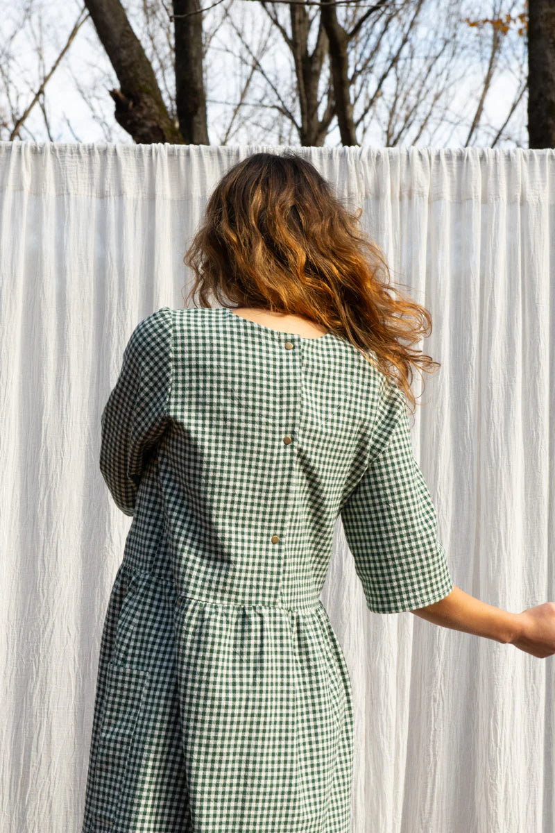 Back view of a woman wearing the Persea Dress by Kazak in Forest Vichy print, a below-the-knee length dress with 3/4 sleeves, a removable belt, and large patch pockets. She is standing in an autumn forest in front of white curtain. 