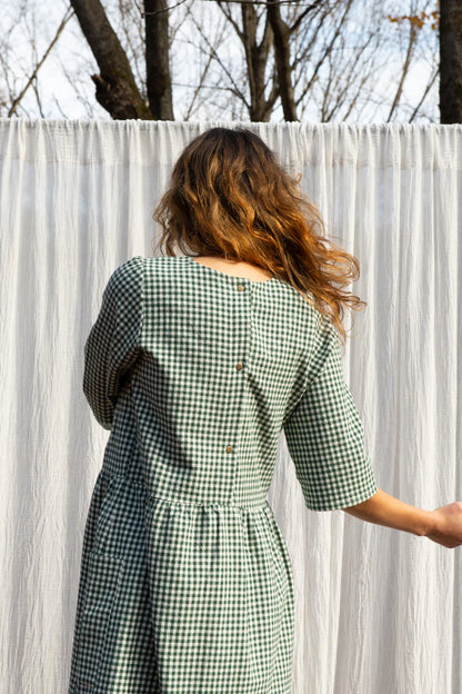 Back view of a woman wearing the Persea Dress by Kazak in Forest Vichy print, a below-the-knee length dress with 3/4 sleeves, a removable belt, and large patch pockets. She is standing in an autumn forest in front of white curtain. 