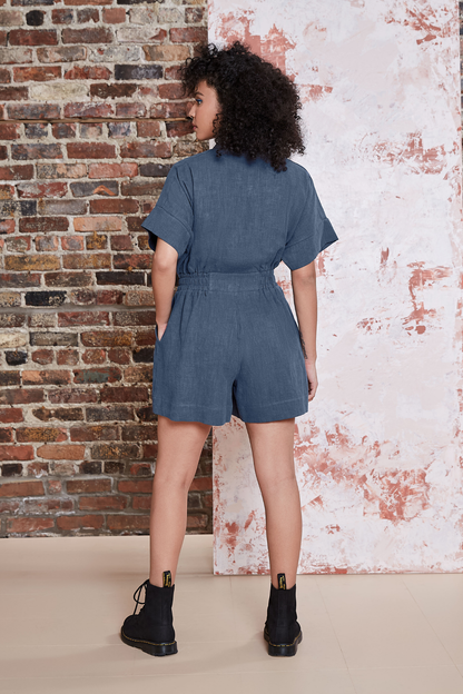 Back view of a woman wearing the Kerouac Jumpsuit by Melow in Slate, a short jumpsuit with a collar, zip front,  chest pockets, elastic waist, and short sleeves. She is standing in front of a brick wall. 