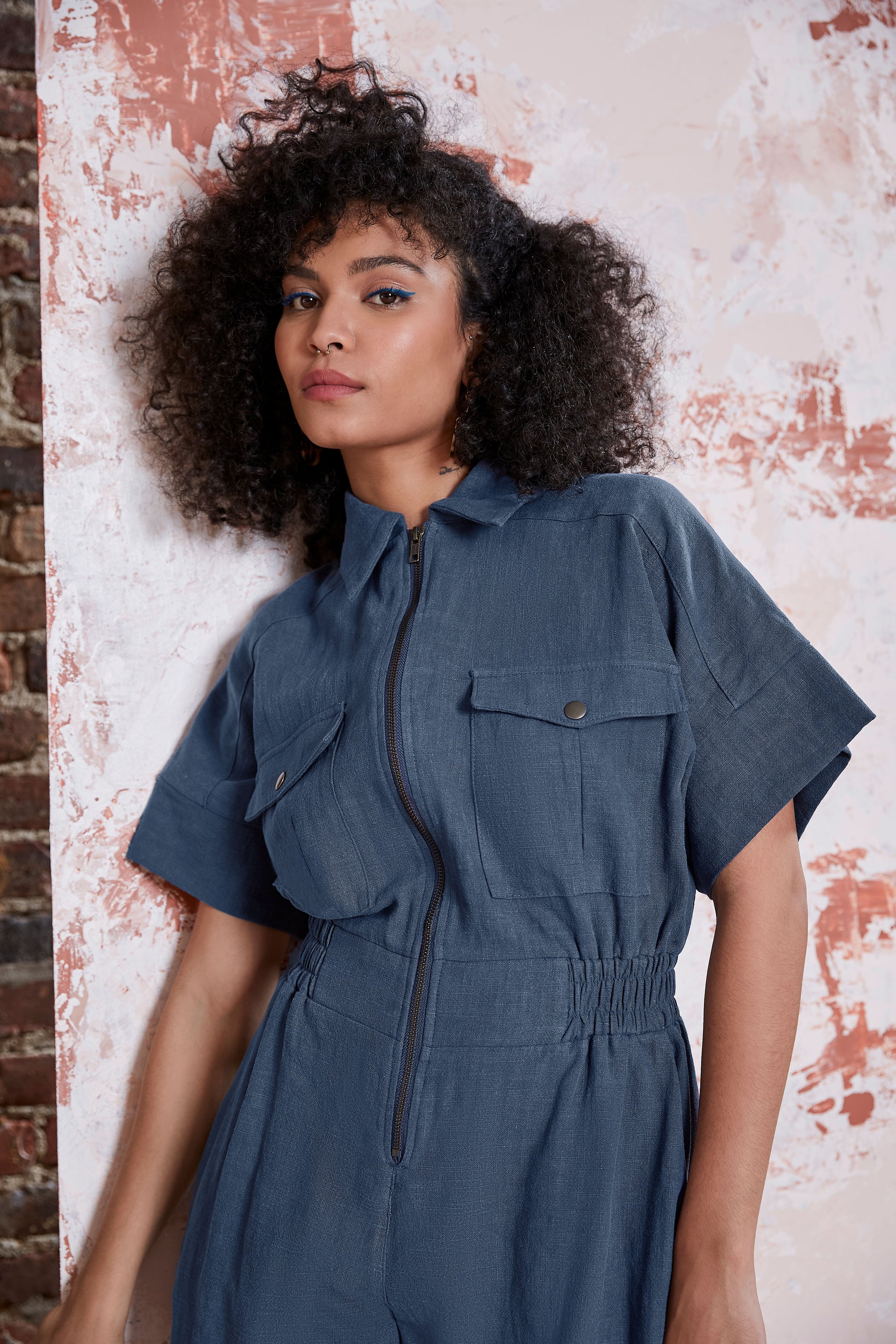 Waist up view of a woman wearing the Kerouac Jumpsuit by Melow in Slate, a short jumpsuit with a collar, zip front, chest pockets, elastic waist, and short sleeves. She is standing in front of a brick wall. 