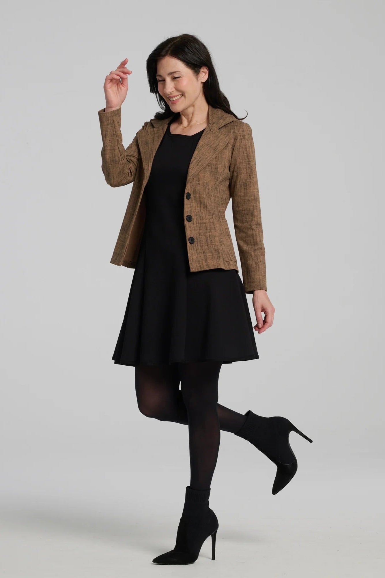 A woman wearing the Toscana Dress by Kollontai in Black, a sleeveless piece with a round neck, princess seams, and fit and flare shape with a full skirt that hits above the knee. She is wearing it with a brown blazer and standing against a grey background.