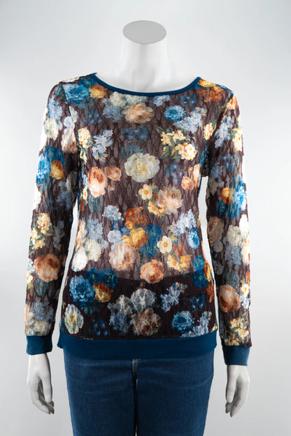 The Aubade Lace Top by Karkass, featuring a floral print on a brown background, round neck, and long sleeves with contrasting teal at the collar, cuffs, and hem, is shown on a mannequin with jeans in front of a white background. 