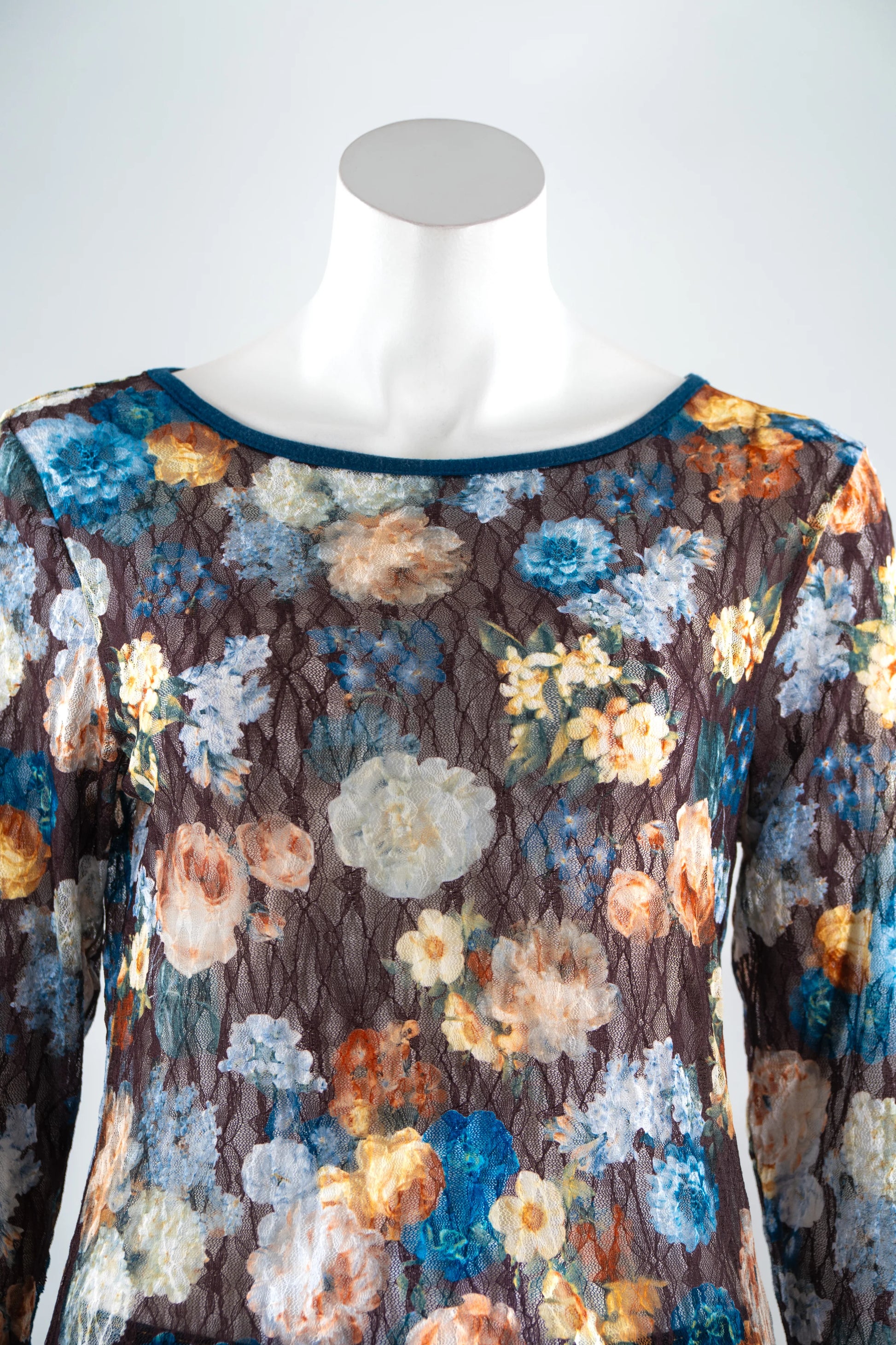 A close up view of the Aubade Lace Top by Karkass, featuring a floral print on a brown background, round neck, and long sleeves with contrasting teal at the collar, cuffs, and hem, is shown on a mannequin front of a white background. 