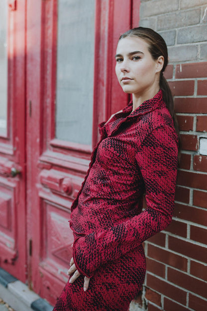 Side view of a woman wearing the Essential Jacket by Karkass in Red Snakeskin, a fitted jacket with a ruffled collar, button front, and frills at the hem and cuffs. She is wearing it with matching pants and leaning against a brick wall. 