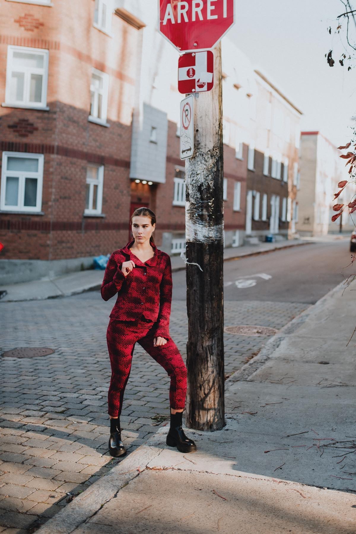 A woman wearing the Essential Jacket by Karkass in Red Snakeskin, a fitted jacket with a ruffled collar, button front, and frills at the hem and cuffs. She is wearing it with matching pants and standing on a street. 
