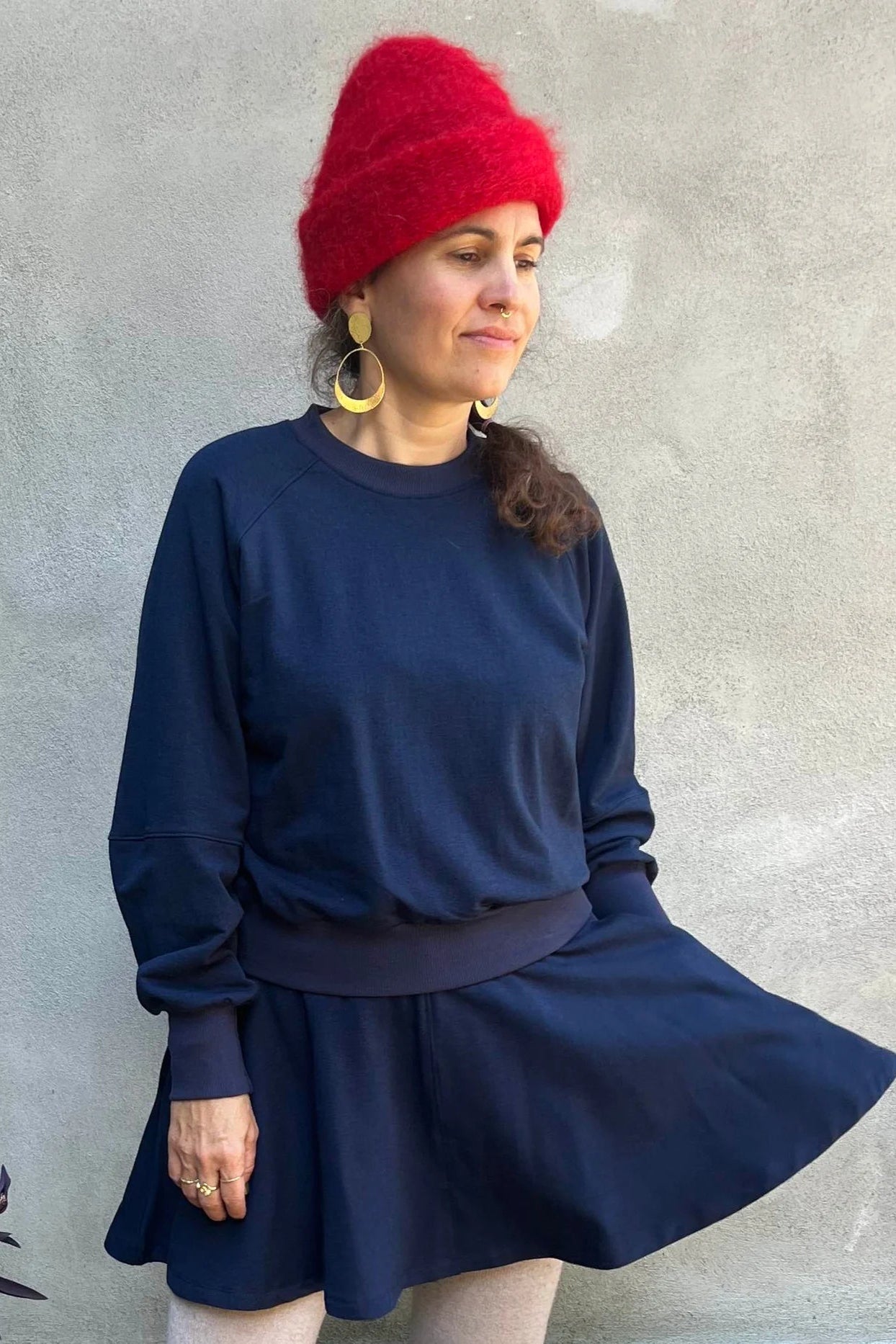 A woman wearing the Juniper Skirt by Kazak in Navy, a full circle skirt with an elastic waist, above the knee length, and side pockets. She is wearing it with matching top and standing in outside in front of a wall. 