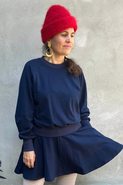 A woman wearing the Juniper Skirt by Kazak in Navy, a full circle skirt with an elastic waist, above the knee length, and side pockets. She is wearing it with matching top and standing in outside in front of a wall. 