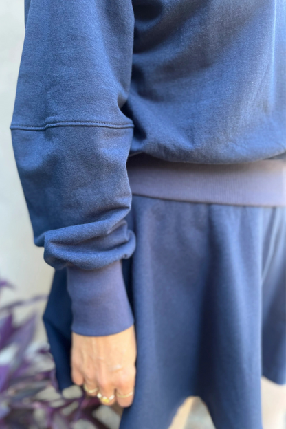 Close up view of a woman wearing the Juniper Skirt by Kazak in Navy, a full circle skirt with an elastic waist, above the knee length, and side pockets. She is wearing it with a matching top and standing in outside in front of a wall. 