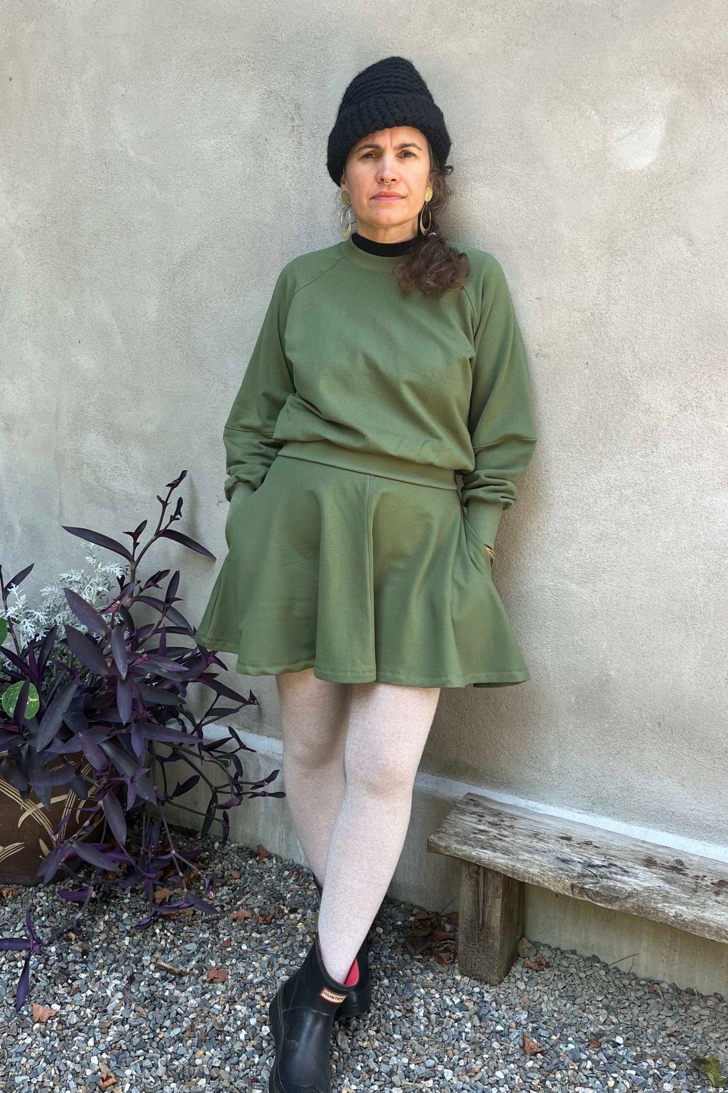 A woman wearing the Juniper Skirt by Kazak in Olive, a full circle skirt with an elastic waist, above the knee length, and side pockets. She is wearing it with a matching top and standing in outside in front of a wall. 