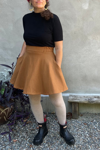 A woman wearing the Juniper Skirt by Kazak in Teak, a full circle skirt with an elastic waist, above the knee length, and side pockets. She is wearing it with a black top and standing in outside in front of a wall. 