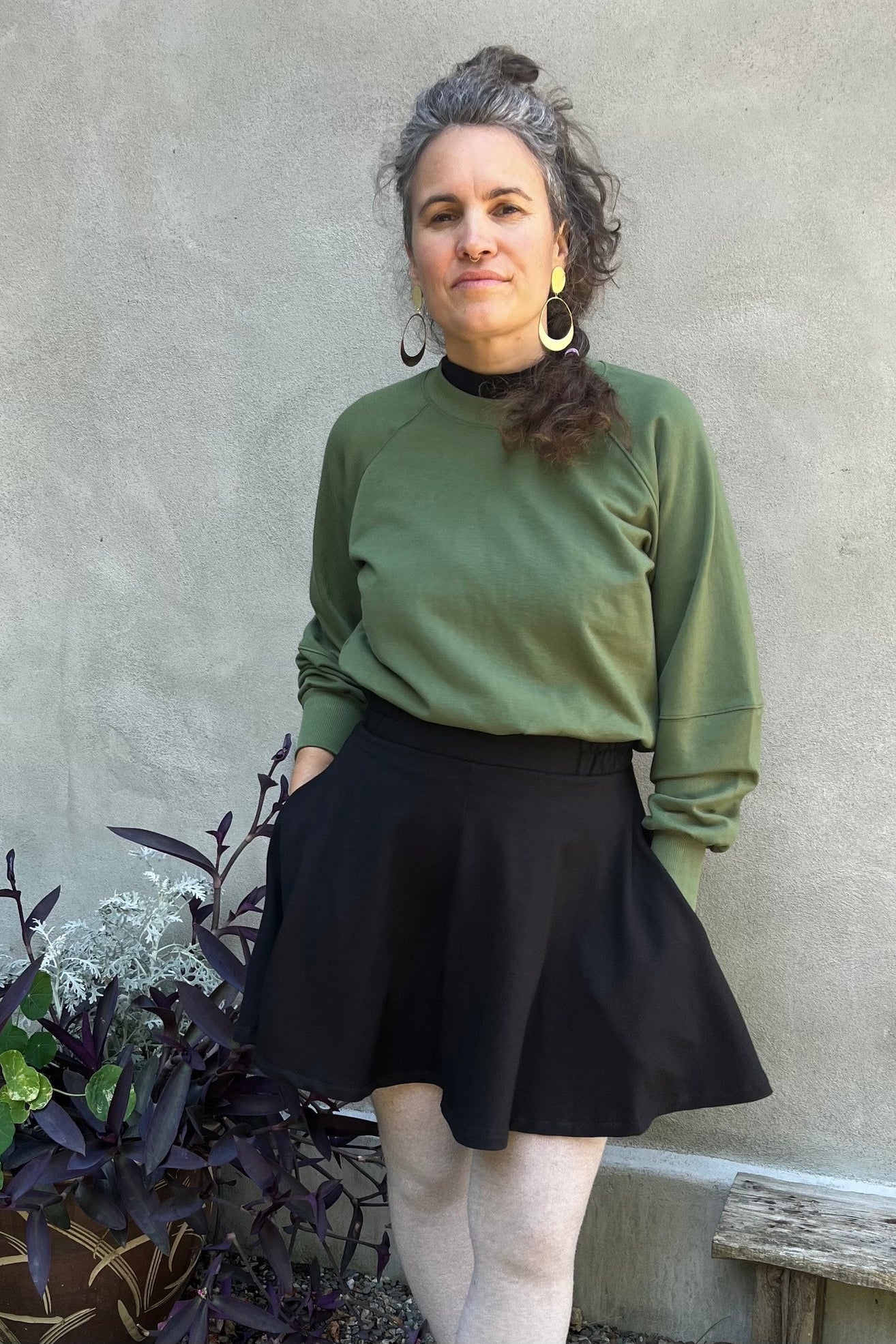 A woman wearing the Juniper Skirt by Kazak in Black, a full circle skirt with an elastic waist, above the knee length, and side pockets. She is wearing it with a the Marilou Crewneck in Olive and standing in outside in front of a wall. 