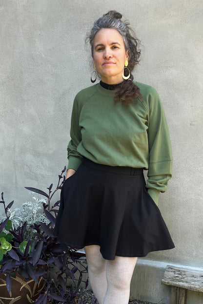 A woman wearing the Juniper Skirt by Kazak in Black, a full circle skirt with an elastic waist, above the knee length, and side pockets. She is wearing it with a the Marilou Crewneck in Olive and standing in outside in front of a wall. 