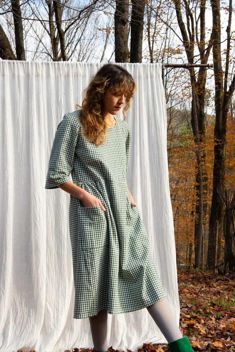 A woman wearing the Persea Dress by Kazak in Forest Vichy print, a below-the-knee length dress with 3/4 sleeves, a removable belt, and large patch pockets. She is standing in a forest in front of a white sheet. 