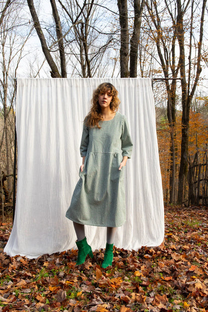 A woman wearing the Persea Dress by Kazak in Forest Vichy print, a below-the-knee length dress with 3/4 sleeves, a removable belt, and large patch pockets. She is standing in a forest against a white background. 
