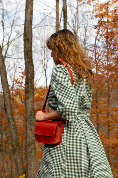 A woman wearing the Persea Dress by Kazak in Forest Vichy print, a below-the-knee length dress with 3/4 sleeves, a removable belt, and large patch pockets. She is walking in an autumn foerst and carrying a red purse. 