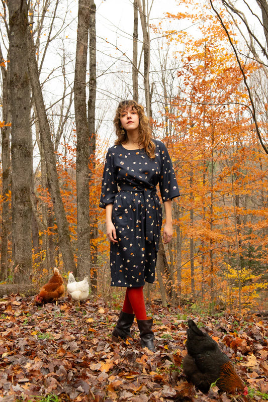 A woman wearing the Persea Dress by Kazak in Midnight Moon print, a below-the-knee length dress with 3/4 sleeves, a removable belt, and large patch pockets. She is standing in an autumn forest with chickens around her.