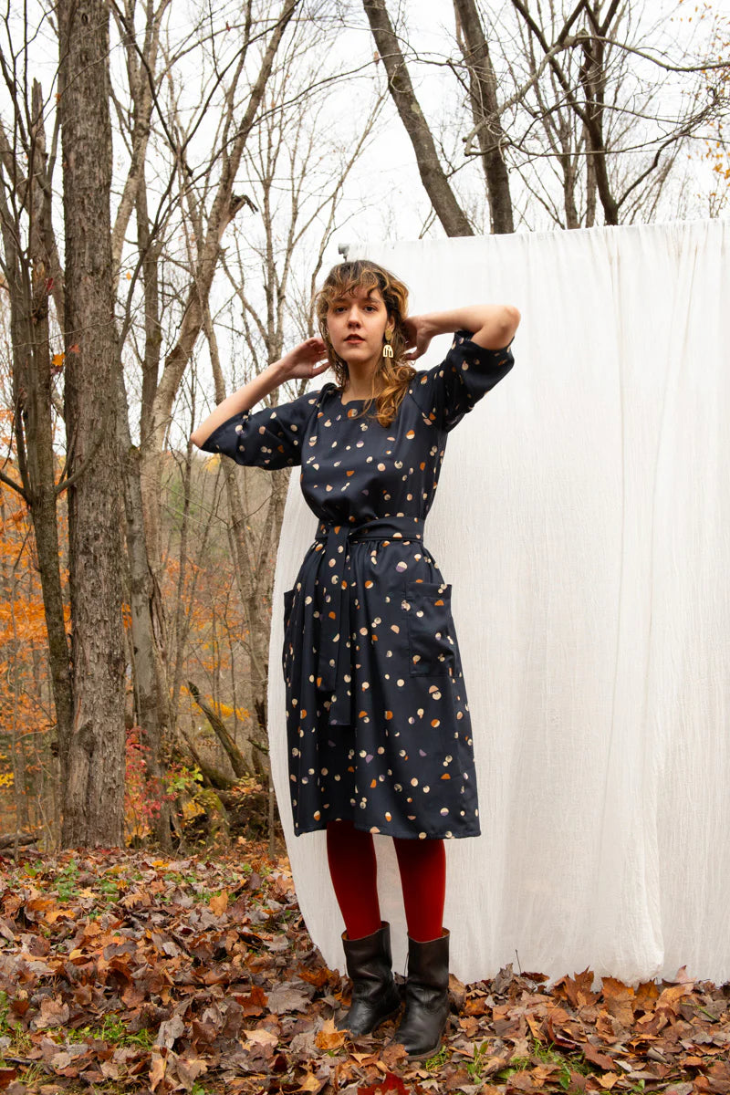A woman wearing the Persea Dress by Kazak in Midnight Moon print, a below-the-knee length dress with 3/4 sleeves, a removable belt, and large patch pockets. She is standing in a forest in front of a white sheet. 