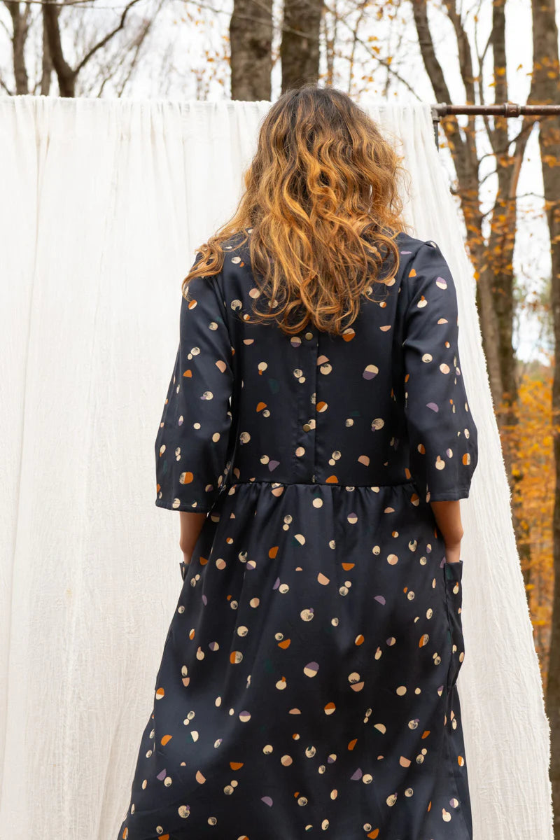 Back view of a woman wearing the Persea Dress by Kazak in Midnight Moon print, a below-the-knee length dress with 3/4 sleeves, a removable belt, and large patch pockets. She is standing in a forest in front of a white sheet. 