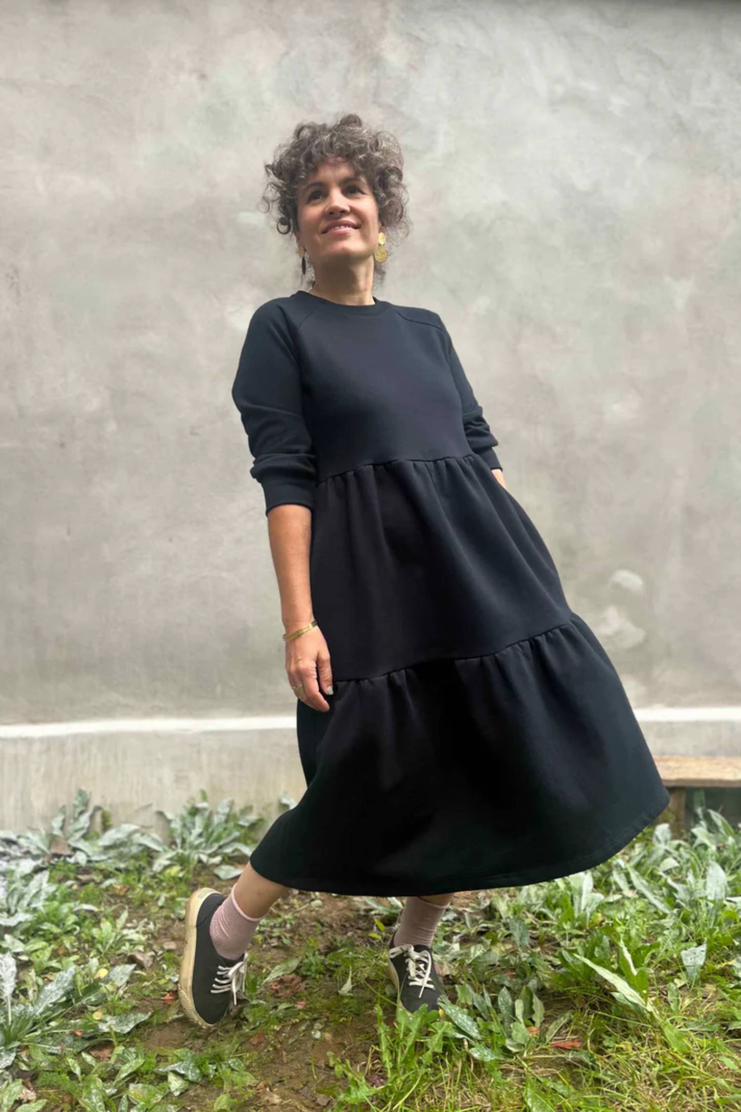 A woman wearing the Simone Dress by Kazak in Black, made from organic cotton and featuring a round neck, 3/4 raglan sleeves, and a tiered midi-length skirt. She is standing outside on front of a grey wall. 
