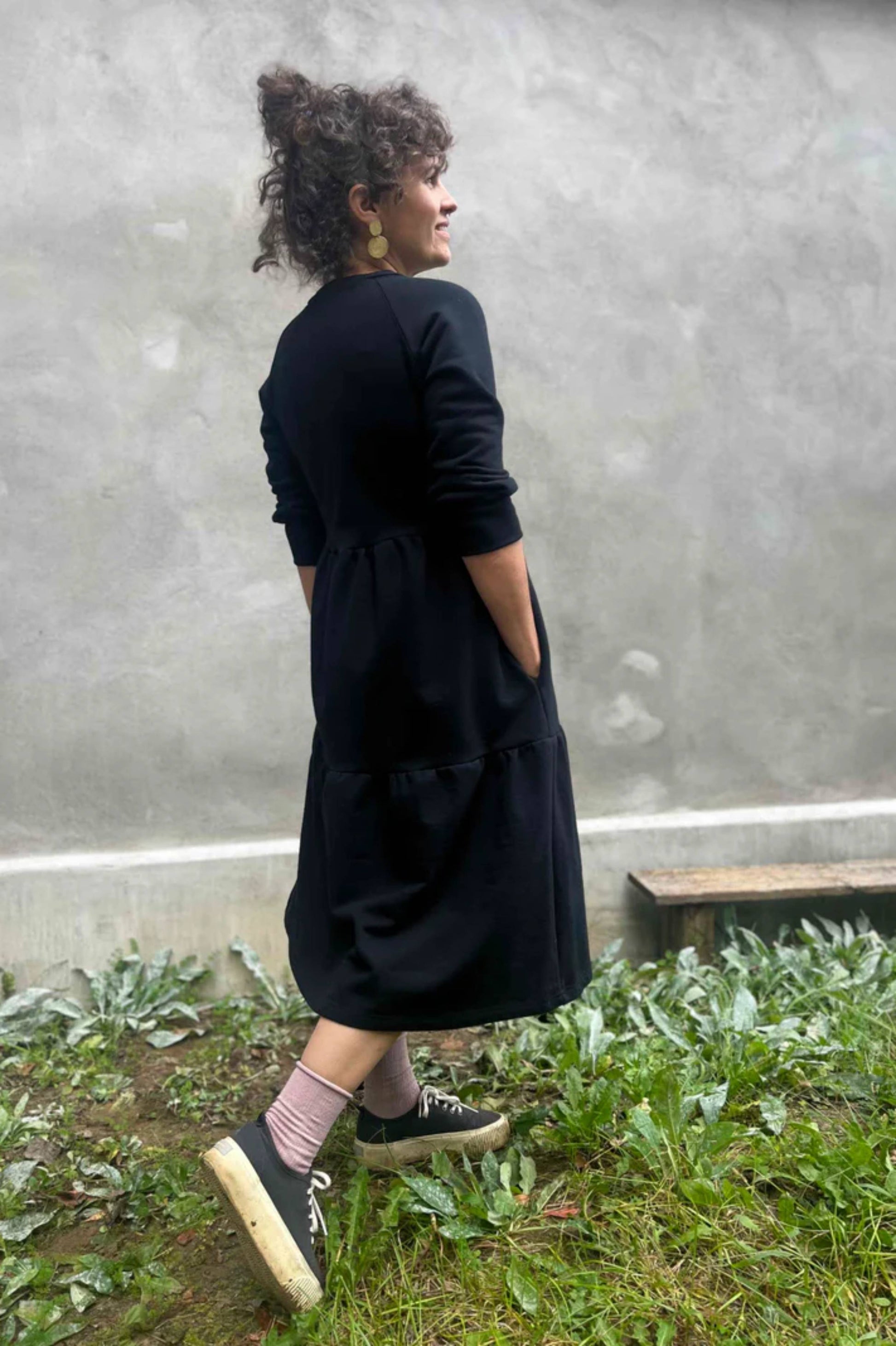 Side view of a woman wearing the Simone Dress by Kazak in Black, made from organic cotton and featuring a round neck, 3/4 raglan sleeves, and a tiered midi-length skirt. She is standing outside on front of a grey wall. 