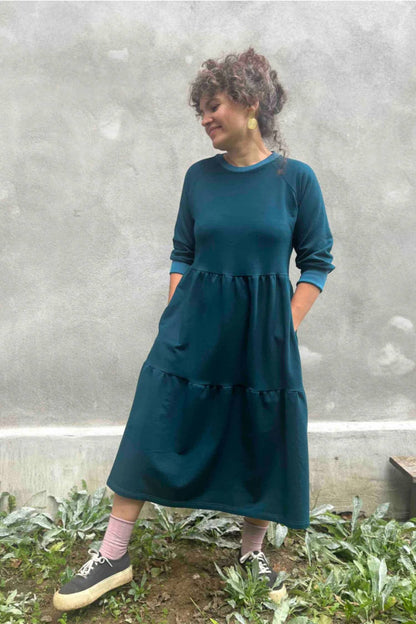 A woman wearing the Simone Dress by Kazak in Teal, made from organic cotton and featuring a round neck, 3/4 raglan sleeves, and a tiered midi-length skirt. She is standing outside on front of a grey wall. 