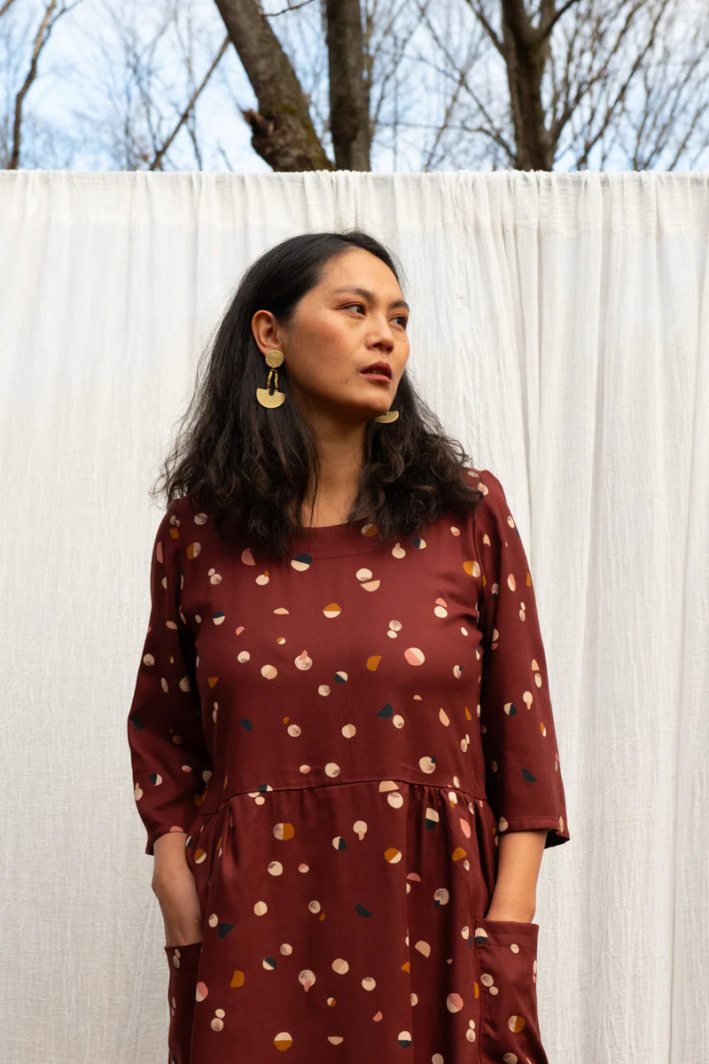 A woman wearing the Persea Dress by Kazak in Burgundy Moons print, a below-the-knee length dress with 3/4 sleeves, a removable belt, and large patch pockets. She is standing in a forest in front of a white sheet. 